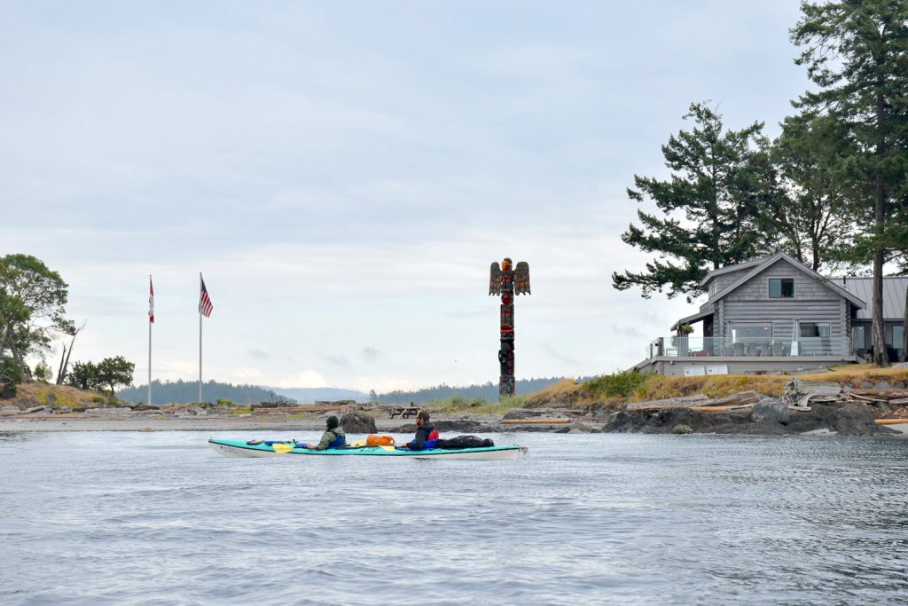 Grey waters at the bottom, Grey sky at the top. There is a strip of land running across the middle of the photo. On the land is a large grey wooden house to the right, totem pole in the middle and two flag poles to the left. A double kayak floats in front of the house as the kayakers look admiringly towards the house.