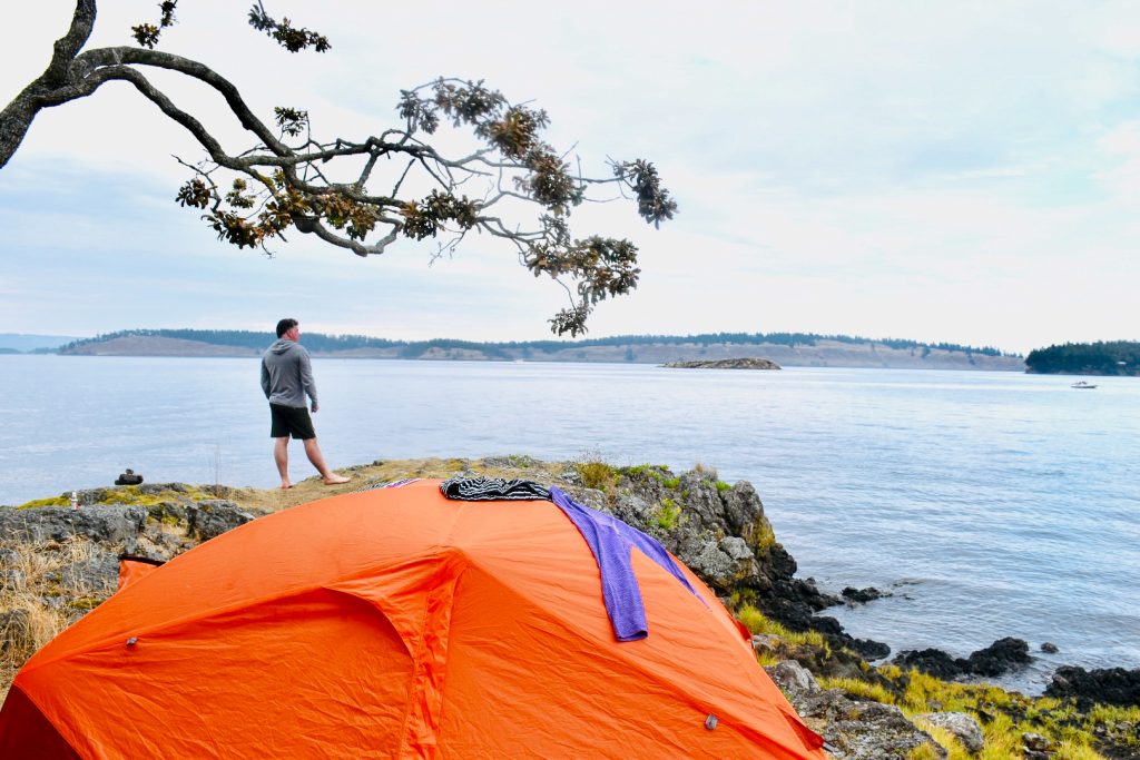 Orange tent in foreground pitched on a rocky shoreline overlooking a flat pale blue sea with land on the horizon and pale grey cloudy sky. A man stands on the rocks looking out over the water. Tree branches coming into the frame from the left. Purple jumper spread out on top of orange tent