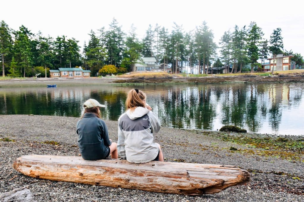 Two young girls sitting on a long log on a pebble beach, looking out over calm water towards wooden houses in fir trees on the horizon.