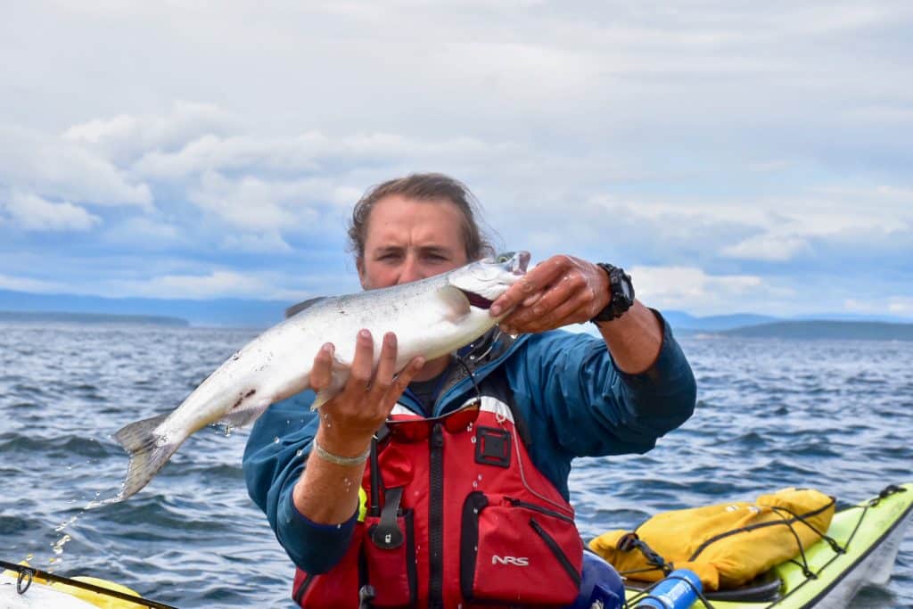 Man in waterproofs holding a large silver fish (salmon) towards the camera. The man is sitting in a yellow kayak on a chopping sea with cloudy sky behind