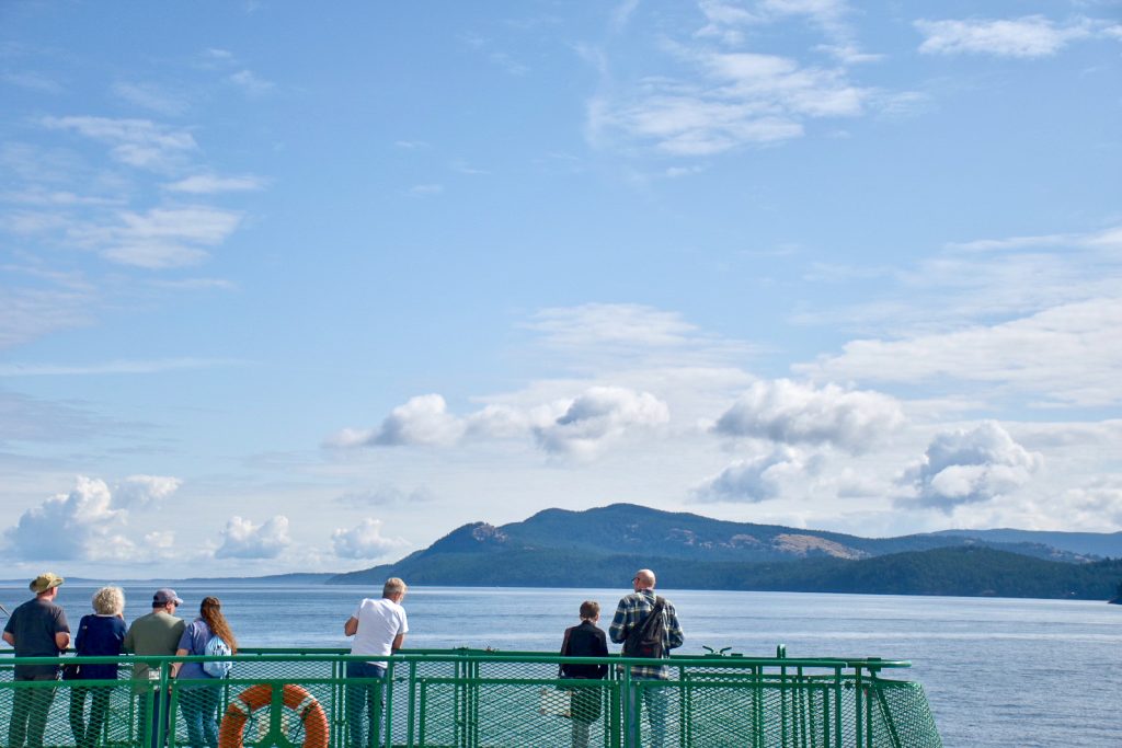 Bottom third of photo features the green railings of a ferry, a lifebuoy and 7 passengers on board looking away from the camera and over a flat body of water behind. Hilly land form on the horizon and blue sky with patchy cloud above