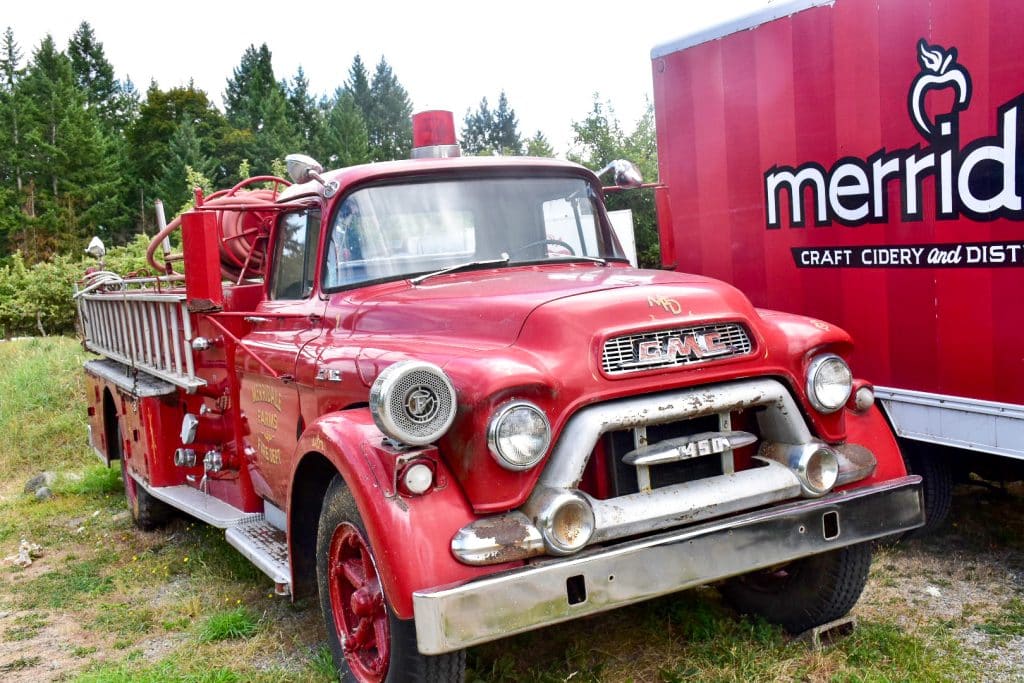 Pale grey sky and forest trees in background. Old fashioned red GMC can in foreground, parked next to a modern lorry with the branding Merridale - Craft Cidery and Distillery
