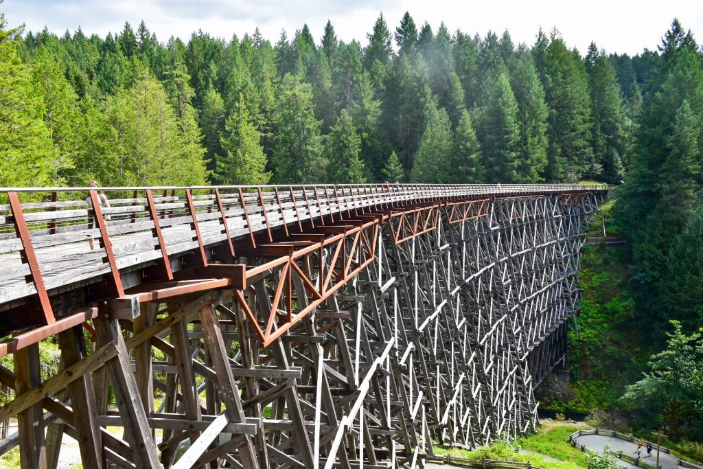 Large wooden lattice bridge (a trestle bridge) running from left to right of frame across a deep valley with forest trees in background