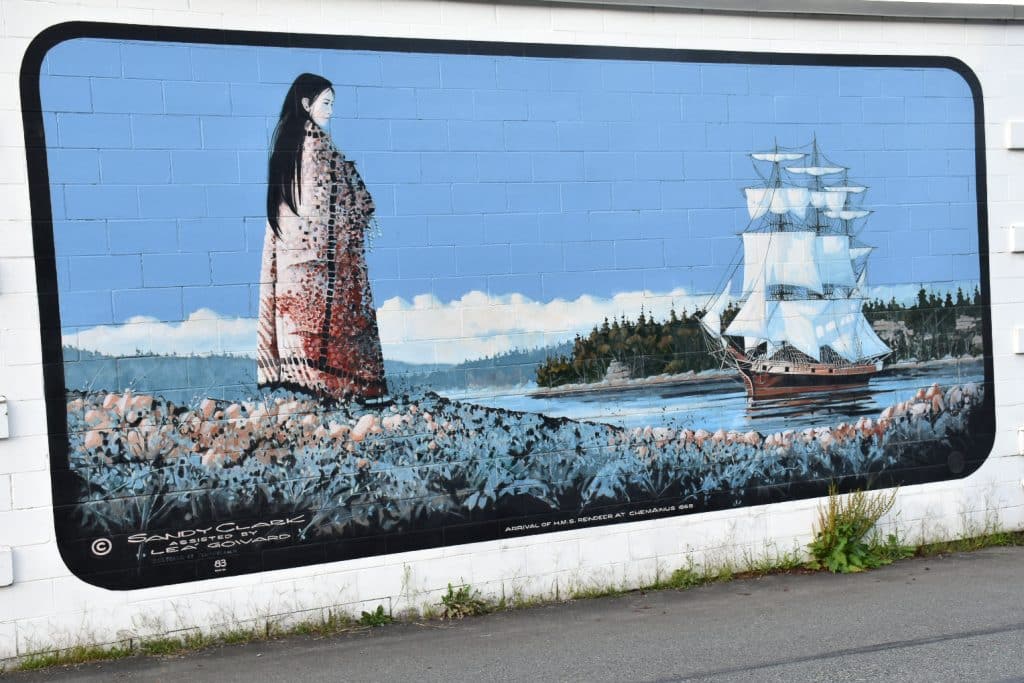 White brick wall with painted mural stretching across photo. The painting has a black border with curved edges. It features a Native American girl with long black hair and long coat looking out across the water to a schooner ship on the water. Fir tree in distance, blue sky and blue ground with pink details