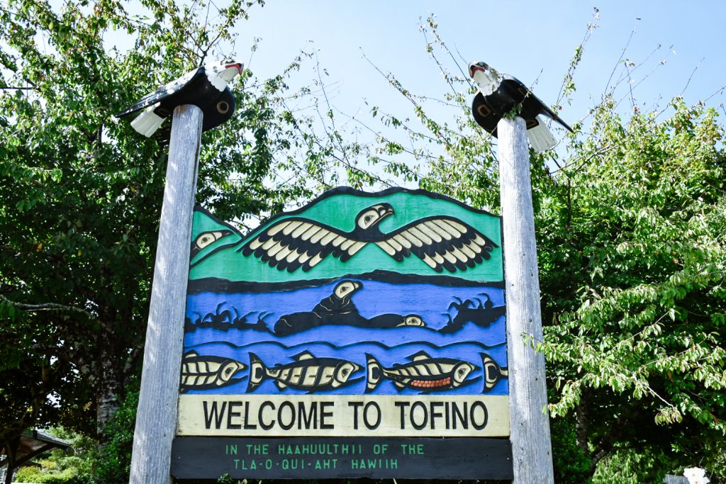 Carved sign saying Welcome to Tofino, featuring two post with carved wooden eagles on the top of each and eagles and fish in the sign itself in green (top) and blue (below). The sign acknowledges the indigenous land on which Tofino stands.