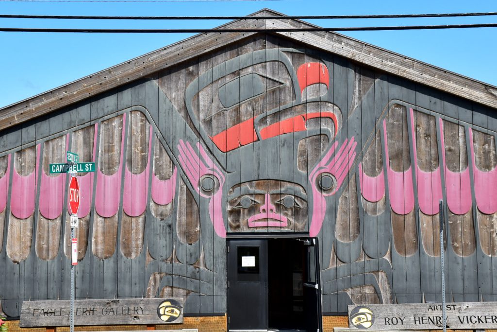 Pale blue sky with a large wooden shed filling most of the frame - the top is an inverted V which is central. There is a doorway with black doors in the middle at bottom of the photo. The walls of the building are decorated with indigenous carvings which look like a thunderbird (with pink and red painted highlights). Sign bottom right says the artist is Roy Henry Vickers. Sign to bottom left says Eagle Erie Gallery