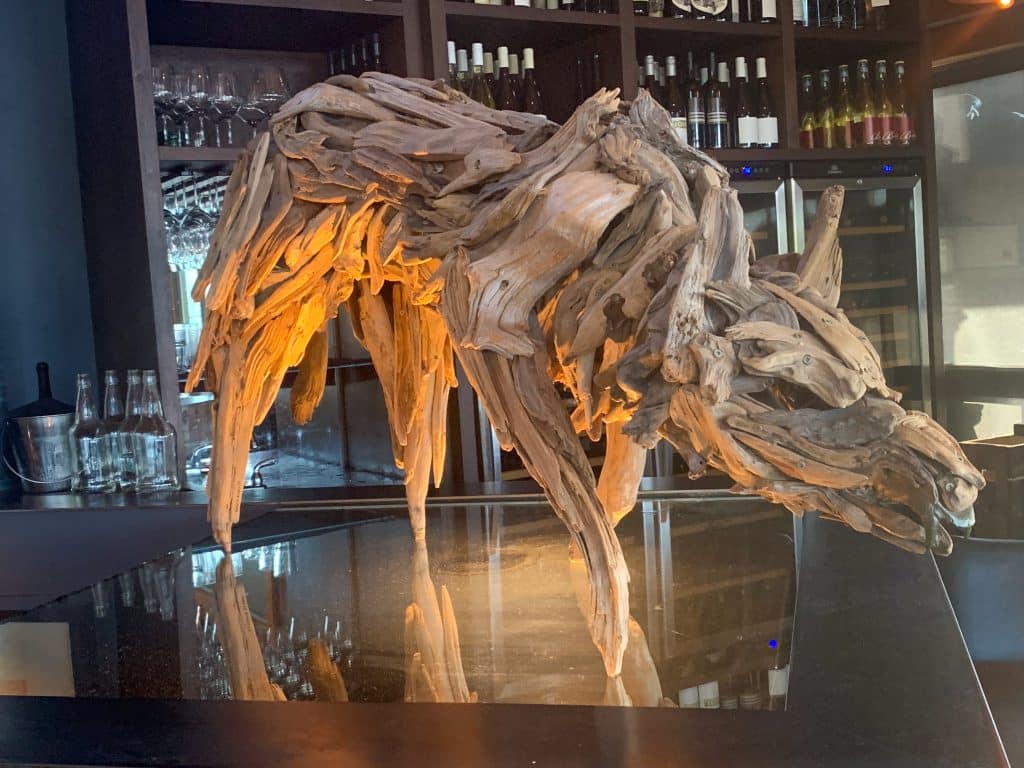 Image taken in a restaurant or bar with shelves of glasses and bottle of wine in the background. In the centre and foreground is a sculpture of a wolf made out of drift wood. The wolf is leaning into the bottom left hand corner of the photo