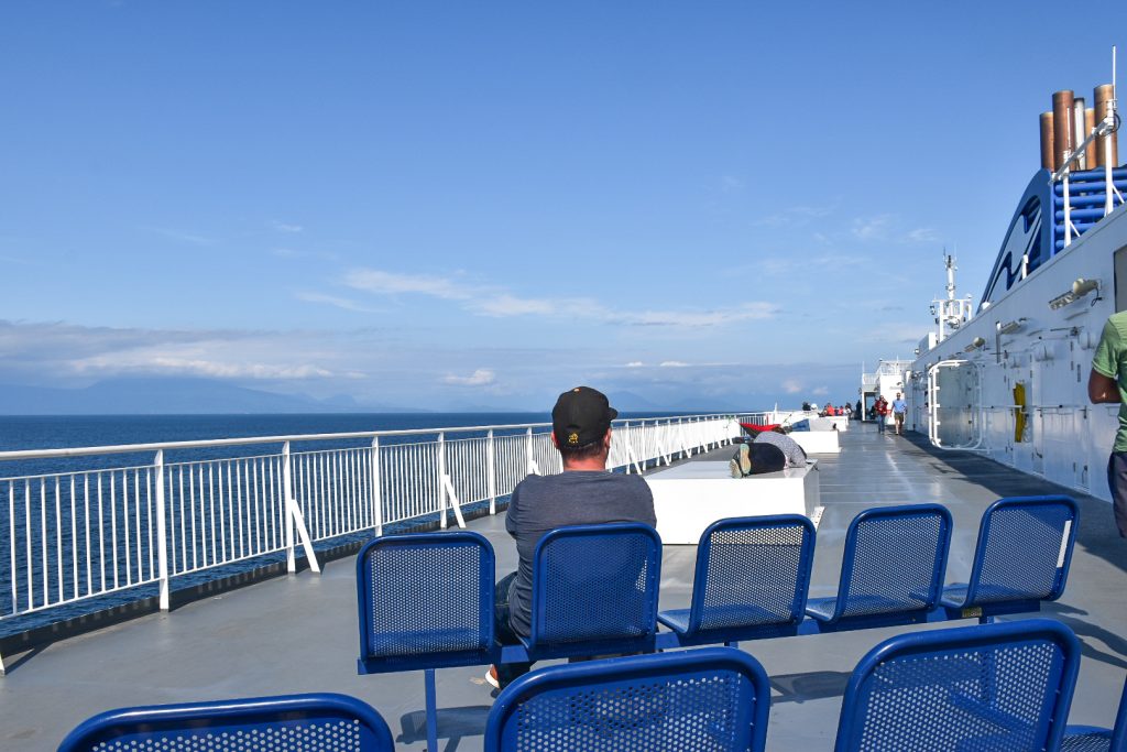 On the outdoor deck of a ferry with white railings, white walls and blue metal chairs. One man in a grey lt-shirt and back baseball hat is sitting with back to the camera looking down the ferry. Blue sea and pale blue sky with faint ships clouds