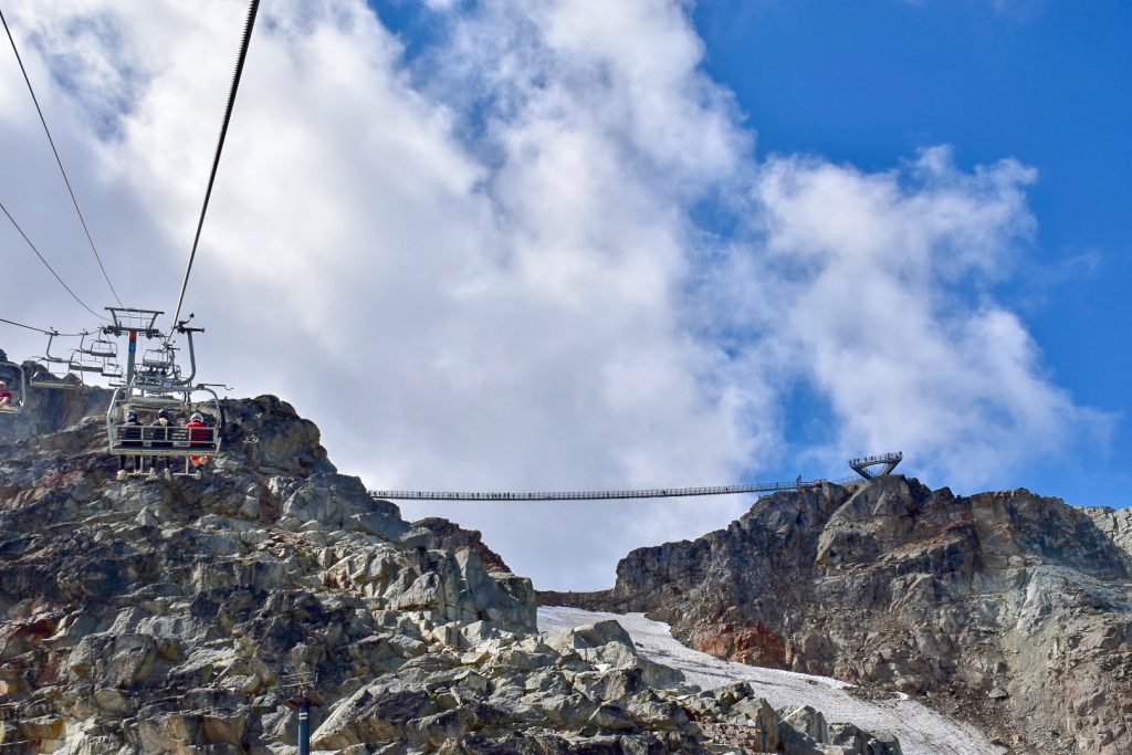 Image looking up a mountain, taken from a chair lift of the other chairs in front to the left of the photo. It is a rocky terrain below with what looks like a patch of snow to the right and blue sky above with large white clouds. You can see a metal bridge spanning a valley in the distance.