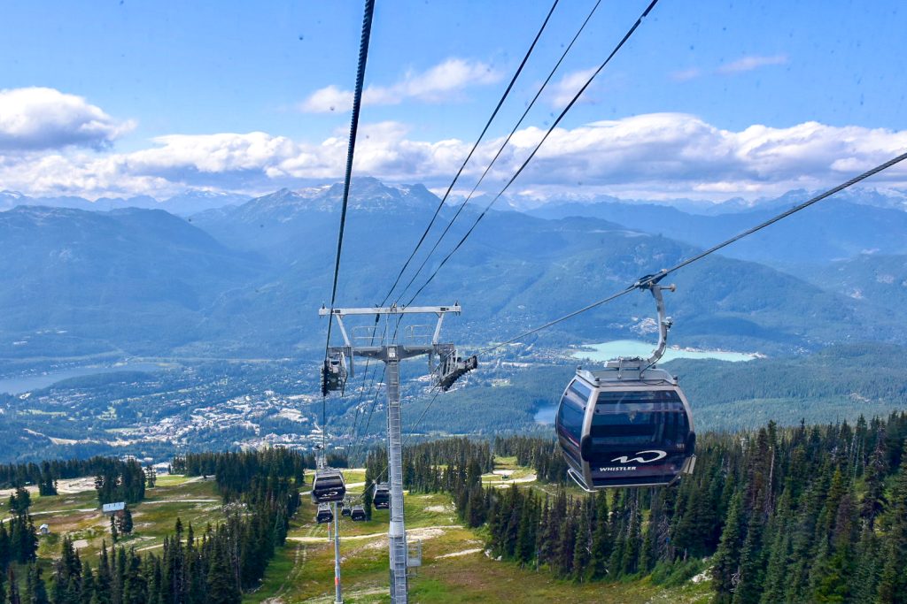 Photo looking down a mountain to valley below. Mountain is green with coverage of fir trees. You can see a village in the base of valley and a couple of lakes. Mountains in distance with clouds. Chair lifts coming up and down the mountain which say Whistler on side