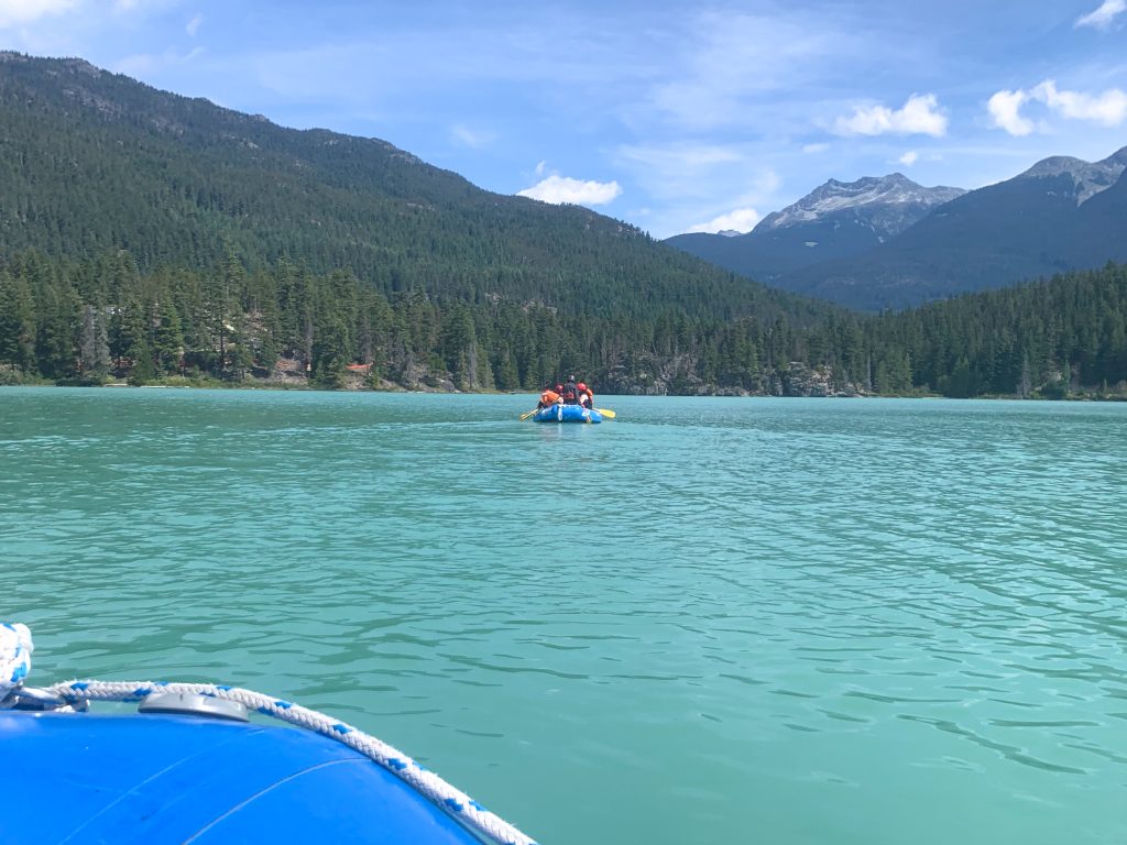 Pale green flat lake with hills in background covered in fir trees, blue sky with some white cloud. ON the water is a blue inflatable raft with crew in lifejackets and yellow oars. Photo taken from a blue raft which is just visible in bottom left of photo