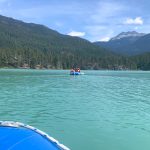 Pale green flat lake with hills in background covered in fir trees, blue sky with some white cloud. ON the water is a blue inflatable raft with crew in lifejackets and yellow oars. Photo taken from a blue raft which is just visible in bottom left of photo
