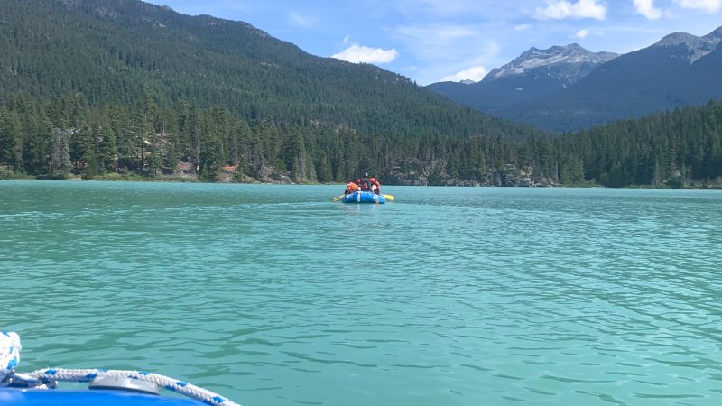 Pale green flat lake with hills in background covered in fir trees, blue sky with some white cloud. ON the water is a blue inflatable raft with crew in lifejackets and yellow oars. Photo taken from a blue raft which is just visible in bottom left of photo