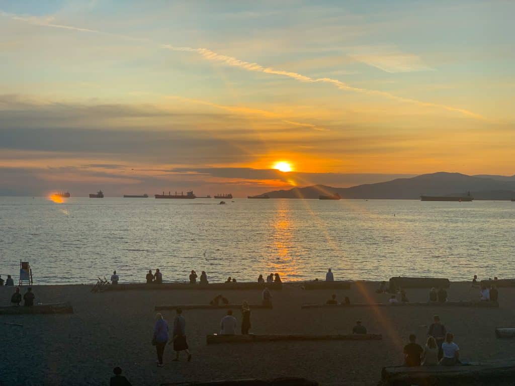 Sunset photo - bottom third is a beach in shadow but you can make out people sitting on logs looking out over the water to the sunset. Middle third is the water. Top third ins the sun setting behind a horizontal whisper of cloud. Golden yellow sky and sun trails