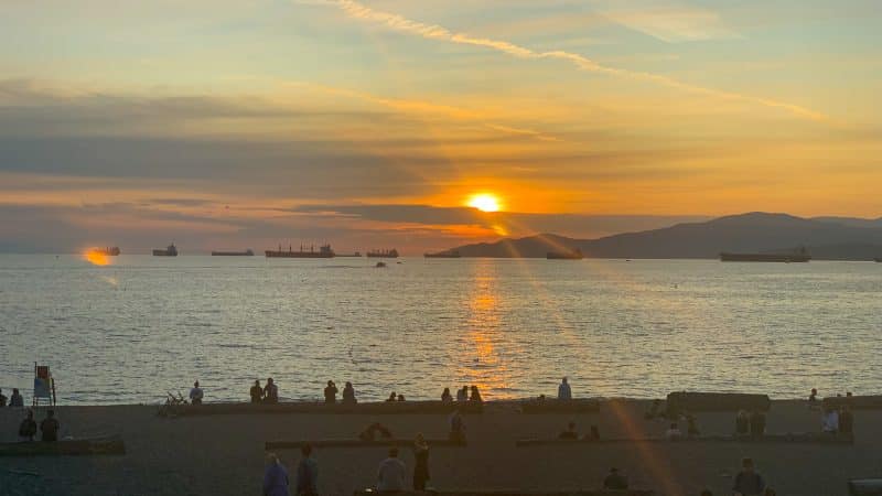 Sunset photo - bottom third is a beach in shadow but you can make out people sitting on logs looking out over the water to the sunset. Middle third is the water. Top third ins the sun setting behind a horizontal whisper of cloud. Golden yellow sky and sun trails