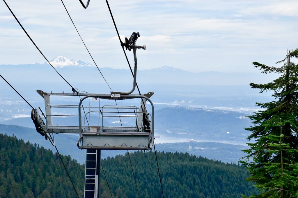 Metal chair left mechanism on left hand side of photo. Chair lift is coming up the hill covered in evergreen trees. Faint outline of snow capped mountain range in background