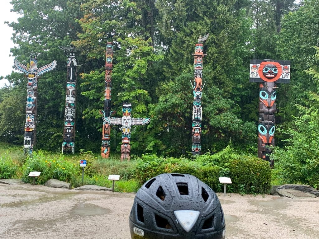 Photo taken from behind someone in a black bicycle helmet looking at a row of multicoloured totems (totem poles) in front of a patch of tall green trees. You can see from the hat and puddles on the ground that it is raining.