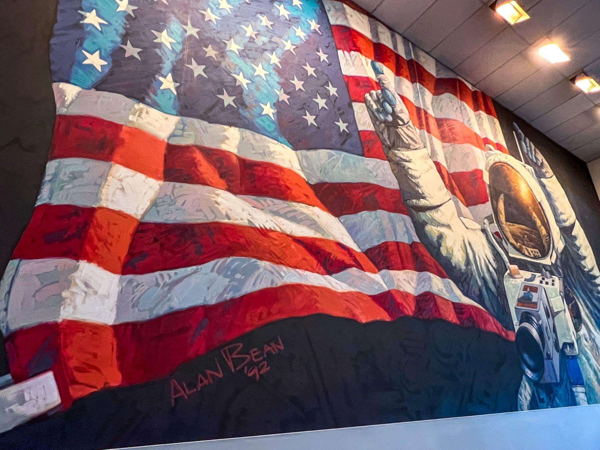 Mural on wall featuring the American Flag billowing towards the camera. On the right hand side of the mural is an Astronaut in white space suit with Gold visor on helmet, with arms in air as if celebrating