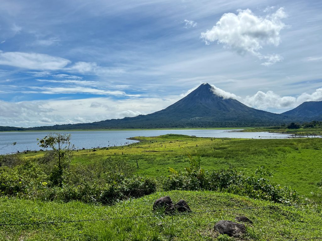 Landscape photo of a conical shaped volcano on the horizon with cloudy blue sky behind and a lake in front of the volcano. Green grassland in the foreground