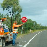 A road with white lines leads from bottom right of frame towards the horizon. On the left of the photo is a workman's van parked by side of the road. A man stands by the side of his van holding a STOP sign (red circle with white letters) to stop traffic. The man is wearing jeans, a high vis jacket over an orange top, Australian flopping had with orange sunshade at the bottom. Trees behind and grey sky above