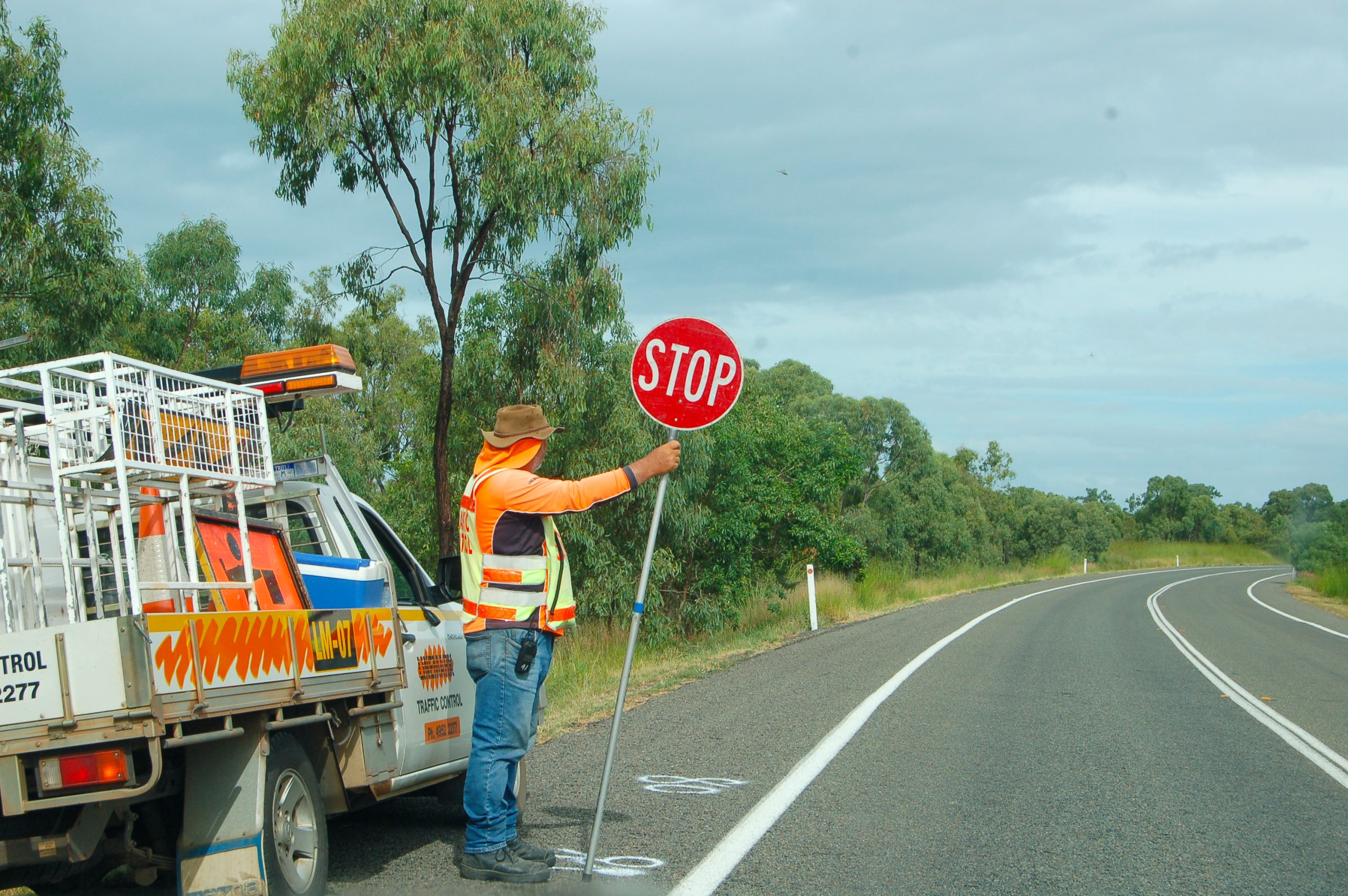 A road with white lines leads from bottom right of frame towards the horizon. On the left of the photo is a workman's van parked by side of the road. A man stands by the side of his van holding a STOP sign (red circle with white letters) to stop traffic. The man is wearing jeans, a high vis jacket over an orange top, Australian flopping had with orange sunshade at the bottom. Trees behind and grey sky above