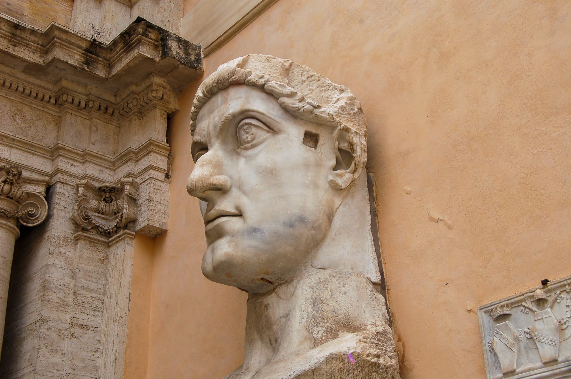 Ocre covered wall with white marble statue of a Roman emperors head against the wall. The head is looking to the left, ou can see the detail on a doorway to the left hand side