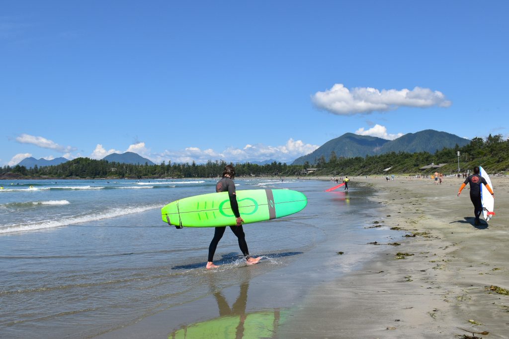 A surfer carrying a large bright green surf board across the centre of the photo, away from the water and towards the sandy beach, the curve of the bay in the background and white surf visible in the water. Trees and mountains on the horizon. Blue sky with just a couple of clouds. Second surfer with white board to the right hand side of frame
