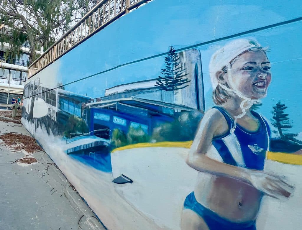 Mural on the side of a building. Right hand side of wall is in foreground. Painting of young girl in navy swim suit and white swim cap, carrying white surf board with yellow trim, running as if to the sea with big smile. Second surfer at the back of the mural