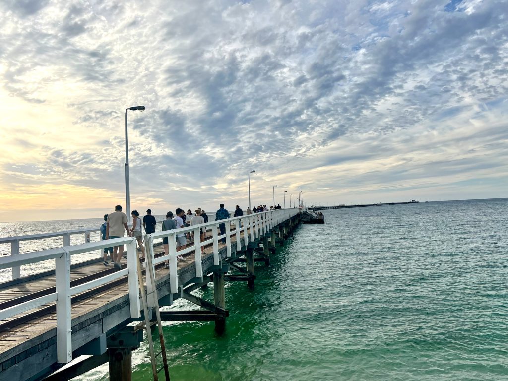 Light is fading over this view of a long wooden jetty with white railings reaching out across a dark green sea. Dramatic clouds in the sky. People are walking along the jeyyu away from the camera
