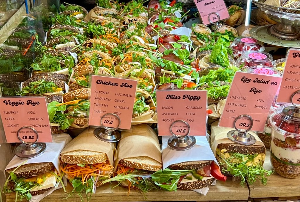 Rows of delicious looking sandwiches lined up in a deli window. The front row have pink signs reading (left to right): Veggie Rye, Chicken Avocado, Miss Piggy, Egg Salad on Rye