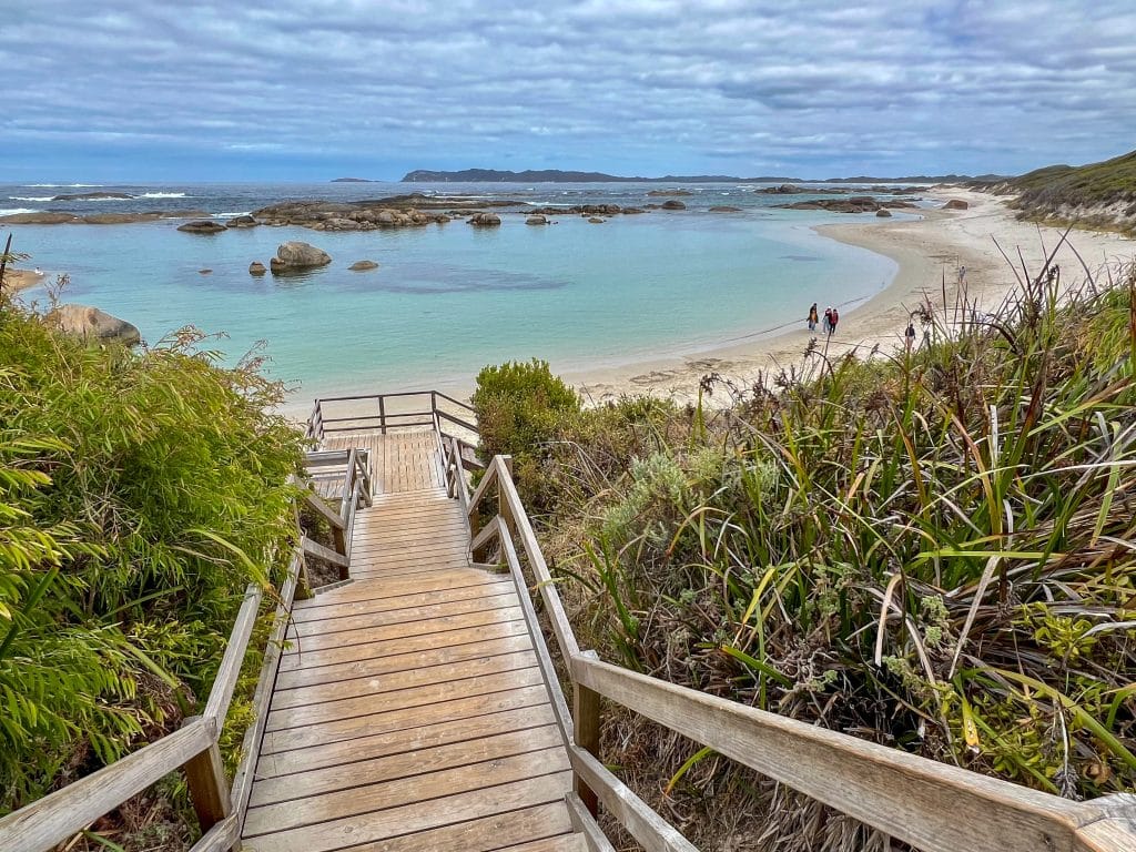 A wooden staircase leads down to a curved sandy beach with rocky barrier on the horizon sheltering s bay of turquoise green water. Cloudy sky