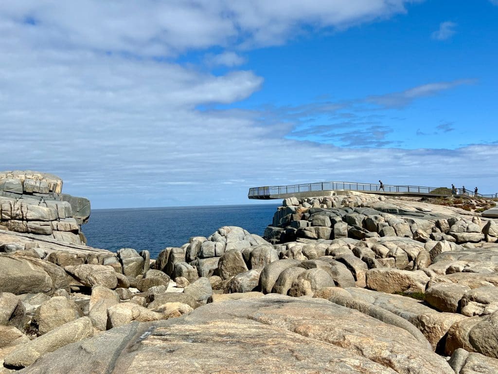Rocky foreground with blue sky above and white cloud to the left hand side. You can see the dark blue ocean through the rocks. There is a metal platform on the rocks to the right hand side, which overhangs the gap. There are 4 people making their way onto the platform