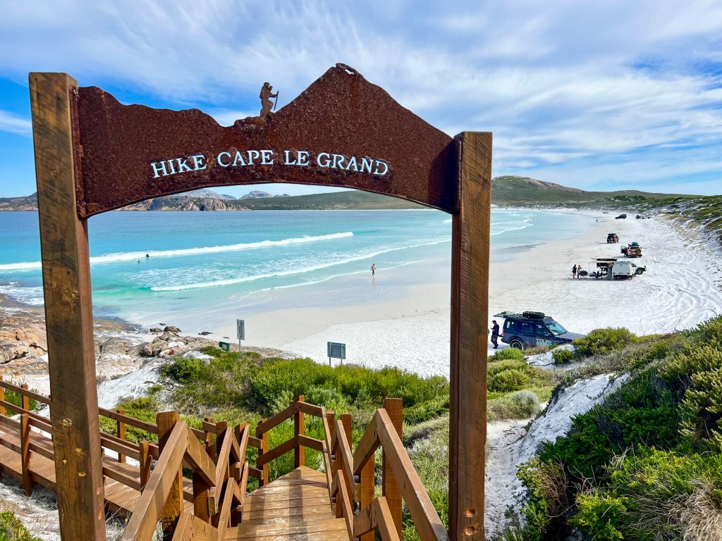 Photo of a curved bay with white sand and pale turquoise waters of the ocean. In the foreground is a wooden staircase leading down to the beach. There is a sign over the staircase which says Hike Cape Le Grand with a hiker carved silhouetted above. Pale blue sky covered with wispy white cloud