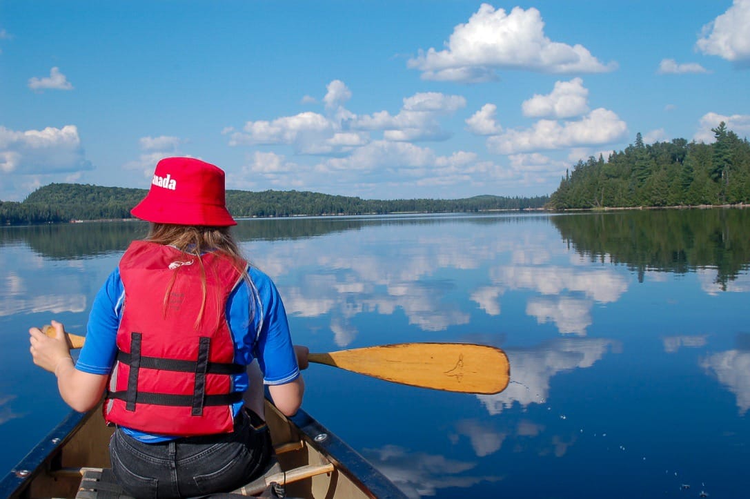 view of a very flat lake, like a mirror featuring the reflections of white fluffy clouds from the blue sky above. There is a girl sitting in a canoe to the left of the picture, She is wearing a blue rash vest, red life jacket and red bucket hat. She is holding a wooden oar out to her right hand side
