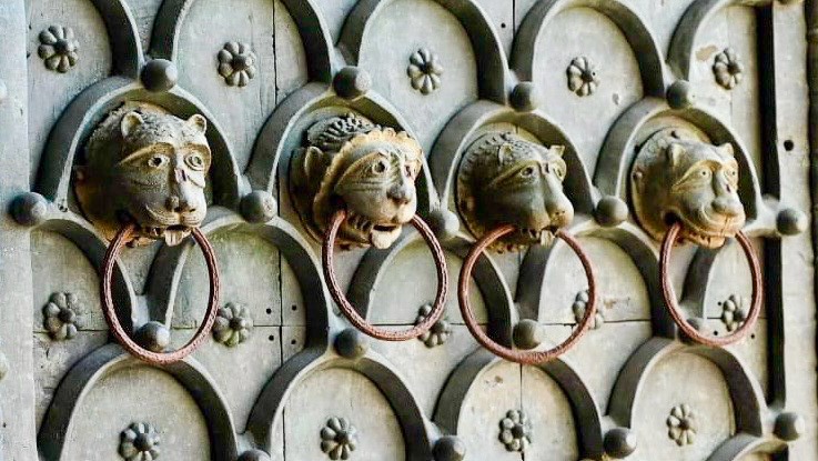 Pale green door carved with arches and flowers. A row of animal heads holding door knockers in their teeth runs across the middle of the photo