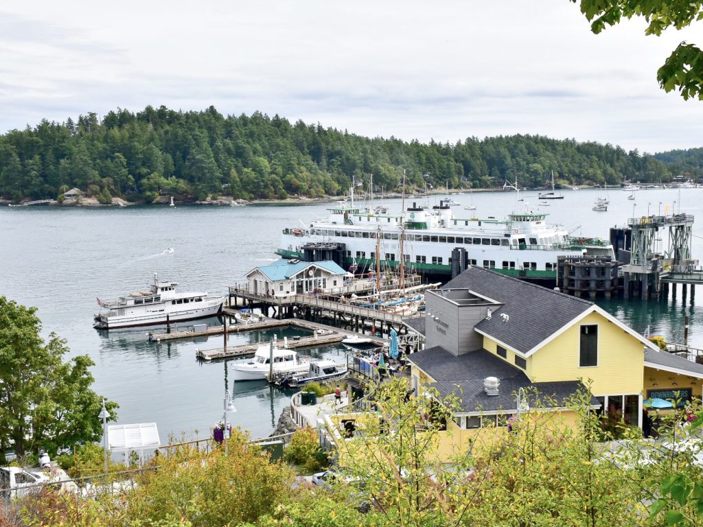 Bottom two-thirds of frame features a small harbour with yellow building, pier, boats moored up and a white and green ferry. Pale grey water in the harbour, green fir trees on horizon and pale grey sky