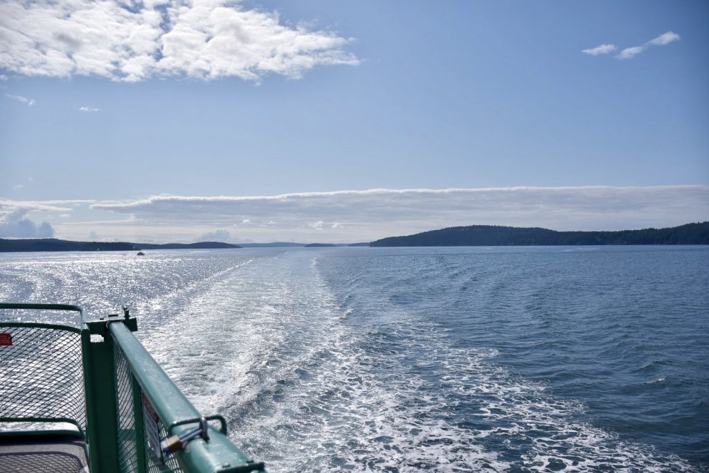 photo of the wake at the back of a boat - small white peaks on an otherwise calm body of water. Green railings of ferry in bottom left of photo. Pale blue sky above with some patches of white cloud
