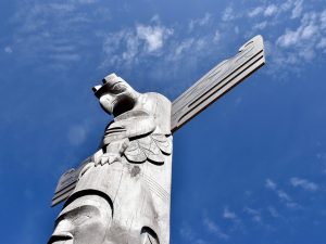 Blue sky with patchy white cloud. A carved grey totem rises up from the bottom left of the frame into the middle of the photo - it as a man's face at the bottom and a thunderbird / eagle at the top with open wings