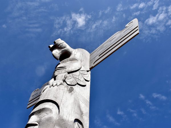 Blue sky with patchy white cloud. A carved grey totem rises up from the bottom left of the frame into the middle of the photo - it as a man's face at the bottom and a thunderbird / eagle at the top with open wings