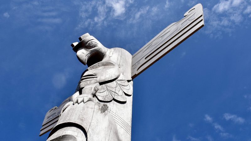 Blue sky with patchy white cloud. A carved grey totem rises up from the bottom left of the frame into the middle of the photo - it as a man's face at the bottom and a thunderbird / eagle at the top with open wings
