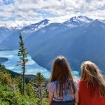 Two girls sit in the foreground looking away from the camera and across a valley filled with snow capped mountains and a turquoise lake in the valley