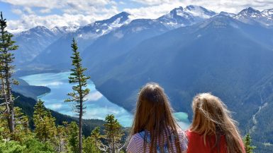 Two girls sit in the foreground looking away from the camera and across a valley filled with snow capped mountains and a turquoise lake in the valley