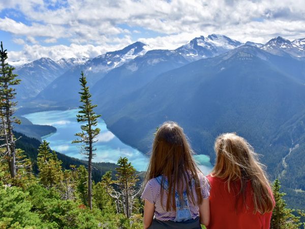 Two girls sit in the foreground looking away from the camera and across a valley filled with snow capped mountains and a turquoise lake in the valley