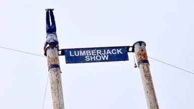 Two wooden posts supported by wires with a blue sign saying Lumberjack Show between them. A man is doing a handstand on the left hand pole.