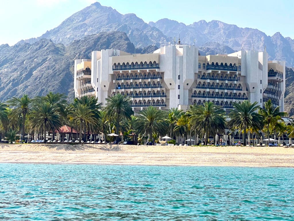 Circular white building with Arabic architectural features behind rows of palm trees and sea. Mountain behind