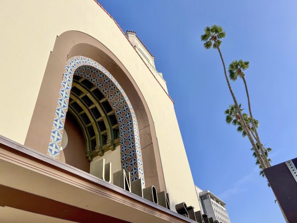 Upwards view of a Spanish Colonial style pale yellow buildings with a large arch and the words Union Station in front. Blue sky to the top right and a couple of palm trees visible