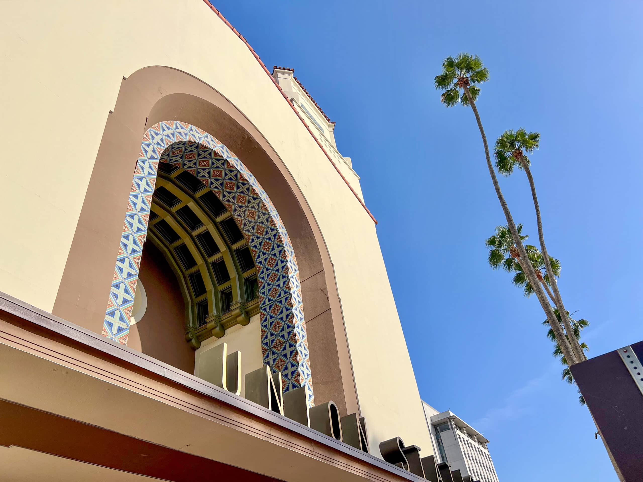 Upwards photo of a cream building with prominent central arch - brown arch, tiled arch and arch with gold and black ceiling. Art Deco signage for Union Station along the bottom. Tall palm trees to the right hand side