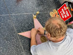 Looking over the shoulder of a man in pale grey t-shirt who is lying on the black pavement engraving a name into a pink star with gold details. Looks like Hollywood Walk of Fame. A redsign to the right says 'Your name in a star'