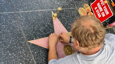 Looking over the shoulder of a man in pale grey t-shirt who is lying on the black pavement engraving a name into a pink star with gold details. Looks like Hollywood Walk of Fame. A redsign to the right says 'Your name in a star'