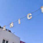 Clear blue sky with two metal wires strung across the space from bottom left to top right. The two wires support old fashioned lightbulb letters spelling 'VENICE". White building to the bottom left